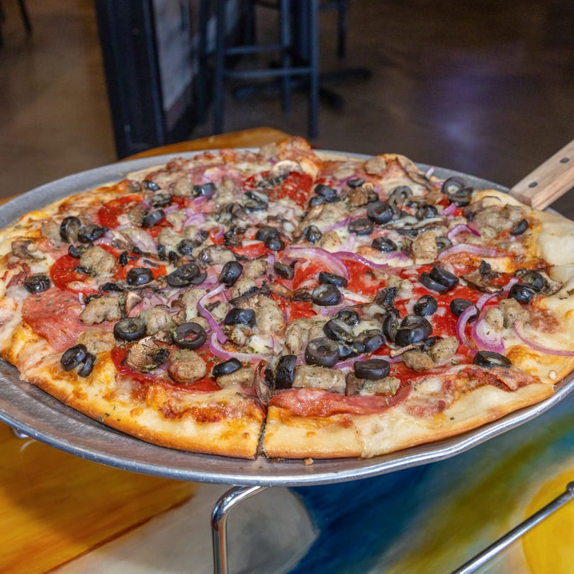A large, freshly baked pizza with pepperoni, sausage, black olives, and red onions on a metal pan.