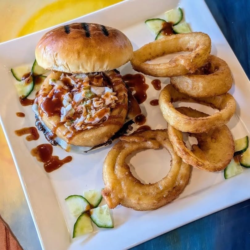A Cold Springs Crossing Bar & Grill burger with onion rings and cucumber slices on a white plate.