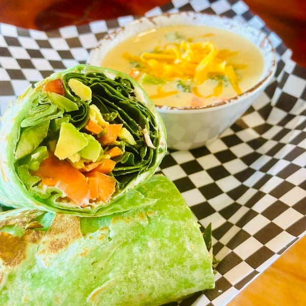 A green wrap filled with lettuce, avocado, and carrots, next to a bowl of cheesy soup.