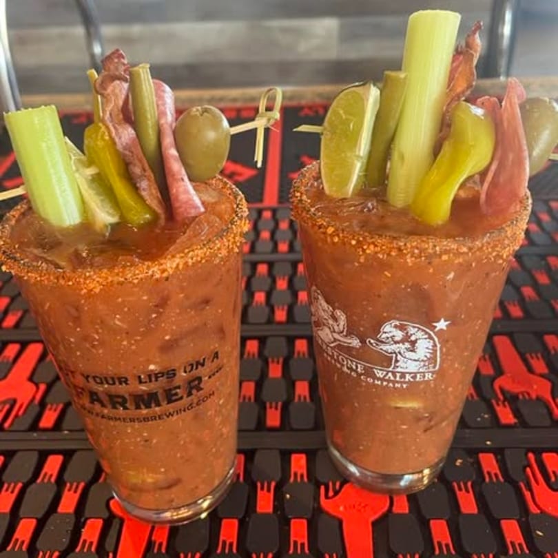 Two Bloody Mary cocktails garnished with celery, limes, olives, and cured meats on a bar mat.