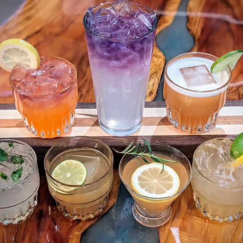 Assortment of six colorful cocktails with various garnishes on a wooden bar top.