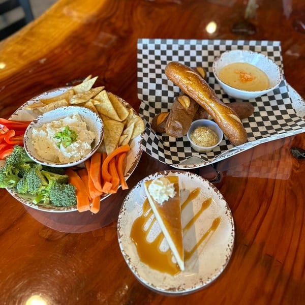 A bar-and-grill spread with pretzel sticks, cheese dip, veggie platter, and cheesecake on a wooden table.