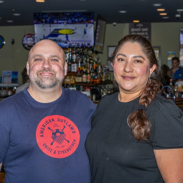 A smiling man and woman stand together in front of a bar with bottles and a TV screen.