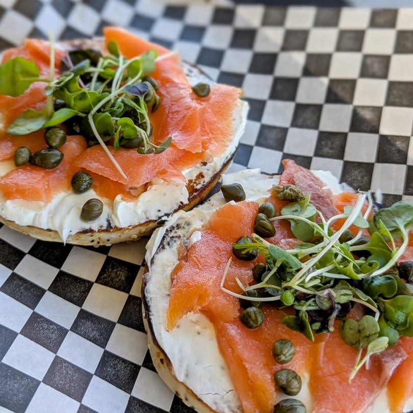 Two bagels with cream cheese, smoked salmon, capers, and microgreens on checkered paper.